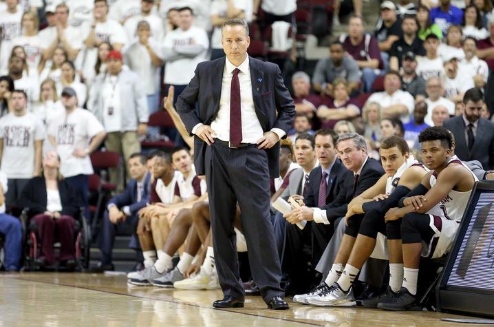 Photo of Billy Kennedy on the sideline of a basketball game. He's wearing a dark suit, white shirt, and maroon tie. His hands are at his hips, and basketball players are seated behind him. 