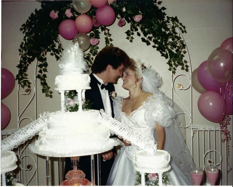Wedding photo of a young white couple. They're standing behind a muiti-tiered wedding cake, with their foreheads touching. An arch of greenery and pink balloons is above them.