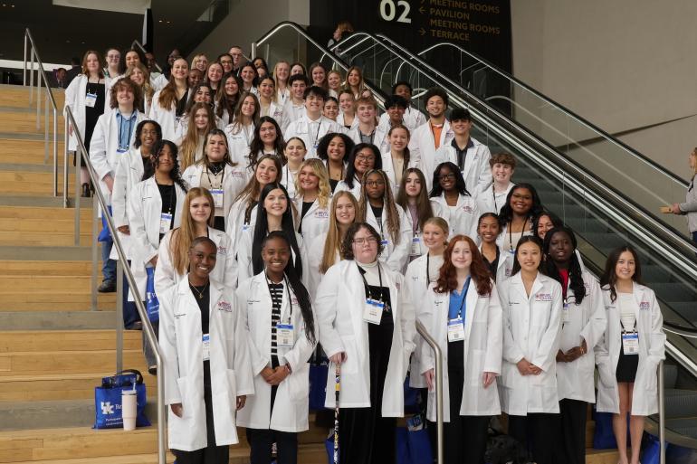 66 high school students in white lab coats stand on a large staircase of a conference center.