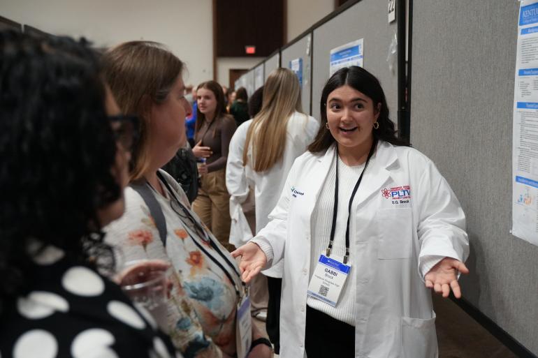 A female student from Frederick Douglass High School, wearing a white lab coat, stands beside her research poster and explains it conference attendees.