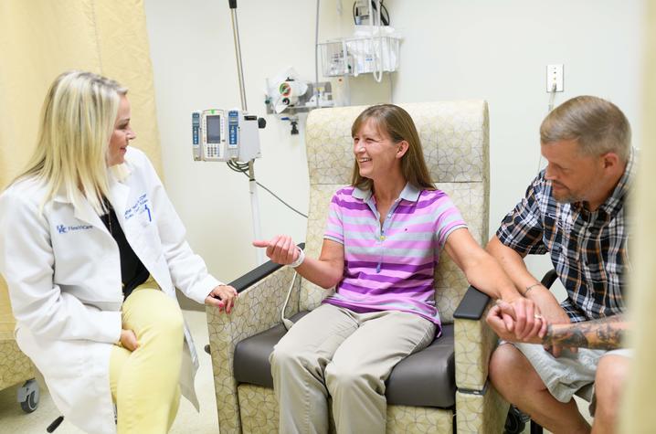 A blond research nurse in a white medical coat sits smiling beside a clinical trial participant who's in a treatment chair. The participant, a middle aged woman with brown hair, is smiling and holding the hand of her husband, who's sitting beside her. Behind her is infusion equipment (IV pole and pump) and medical equipment.