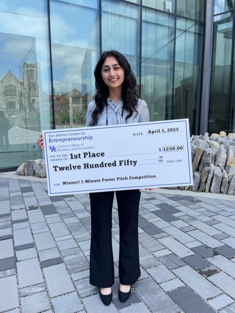 A young woman with long, wavy, dark hair stands holding a giant ceremonial check that says "First Place: $1250". She's standing outside on a paved walkway with a glass building behind her. 