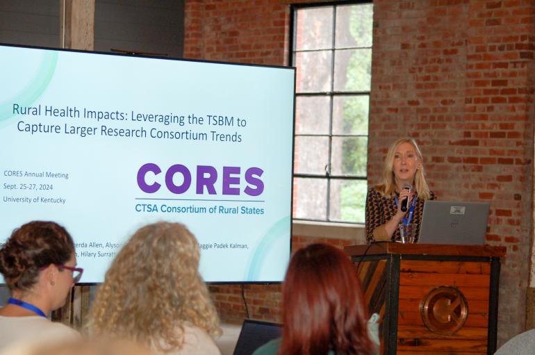 A blond woman stands at a podium, giving a presentation next to a large screen. 