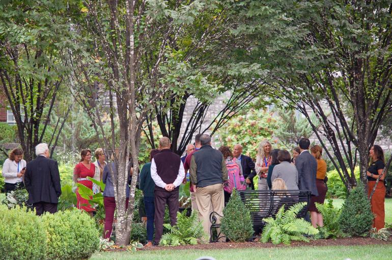 A group of people mingle in a garden under large trees. 