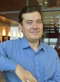 Photo of a middle aged white man which dark brown hair brushed back from his forehead, smiling at the camera. He's wearing a blue collared shirt and his elbow is leaning on something out of the frame. 