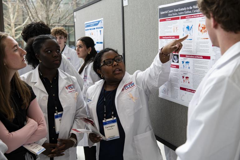 High school students in white lab coats gather around a research poster; one of them is pointing at the poster and speaking. 