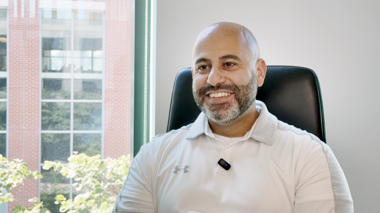 A smiling man sitting in a office chair; he's wearing a white polo shirt and has a bald head and light beard. Behind him is a white wall and a window. 