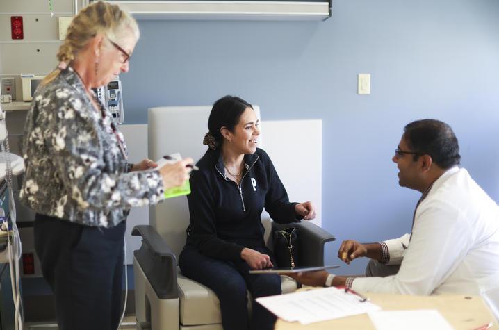 A young woman sits in a hospital chair, smiling at a doctor in a white coat sitting beside her. To the left, a standing woman reviews information on a clipboard. 