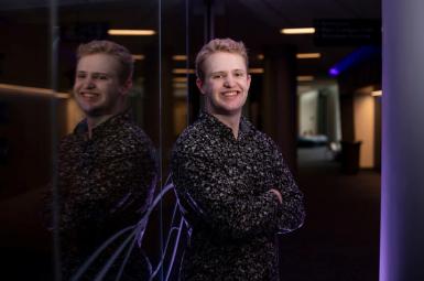 Roberto Obregon Garcia, an undergraduate white male with short blond hair, smiles and leans against a reflective wall. He's wearing a black, patterned button-down shirt and his arms are crossed at his chest. 