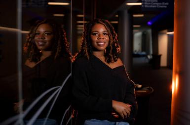 Alexis James, a Black female undergraduate with shoulder length curly hair, learns against a reflective wall. She is smiling, her arms are folding at her hips, and she's wearing a black sweater and jeans. 