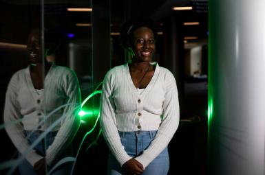 Hope Makumbi, a female, Black undergraduate, smiles and leans against a reflective wall. She's wearing a white button up sweater and jeans, and her short hair is behind a headband. 