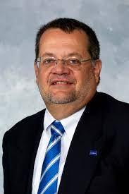 Headshot of Craig Rush, PhD. He's a white, middle-aged man with dark, short-clipped hair, and he's wearing rimless glasses, a dark suit, white collared shirt, and royal blue tie with thin, white, diagonal stripes. 