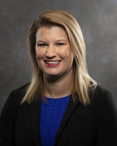 Mid headshot of Ella Dunbar, a white woman with dark blonde, straight hair just past her shoulders. She's wearing a black blazer and a royal blue shirt and she's smiling at the camera. 