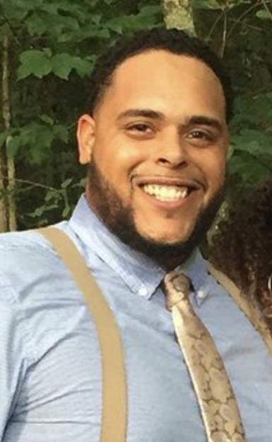 Mid-shot of Robert Bell, a Black man with a broad smile. He's wearing a light blue button-up shirt, tan suspenders, and a light gold tie, standing in front of trees. 