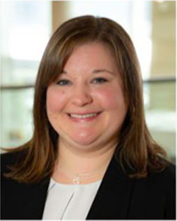 Headshot of Katie Thomas, a white woman in her thirties. She has straight, brown, shoulder-length hair and is wearing a black blazer and a white shirt. She's smiling at the camera. 