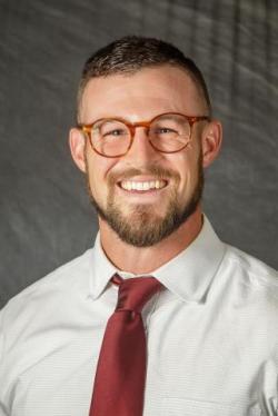 Headshot of Alex Elswick, a white man with short brown hair, a short beard and trimmed mustache. He's smiling at the camera, wearing tortoise shell glasses, a white collared shirt, and a burgandy tie. 