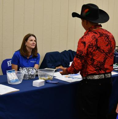 A female CCTS staff member sits behind a table draped in a blue table cloth, talking to a Latino man who is standing across the table from her wearing a red collared shirt, black jeans, and black cowboy hat. 