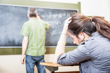 Teenage Girl sitting at classroom desk; struggling to focus on teacher in front of the classroom