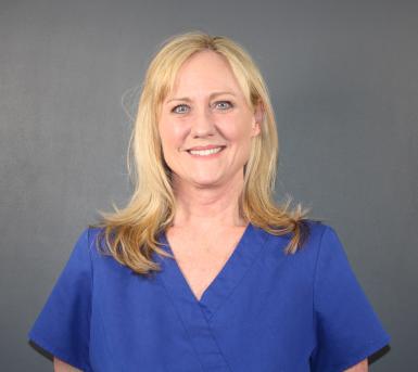 Photo of Alayne Nieto. She's a white woman with blonde hair past her shoulders. She's smiling at the camera, wearing blue scrubs, and standing in front of a gray background. 
