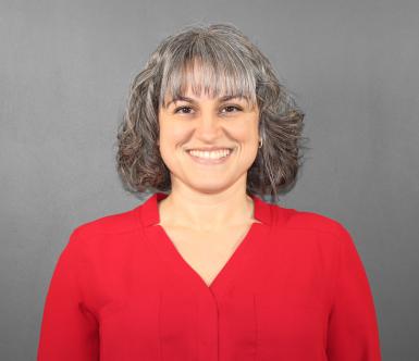 Photo of Shannon Samaha. She has brown-gray hair in a bob just above her shoulders with bangs. She's smiling at the camera, wearing a red shirt, in front of a gray background. 