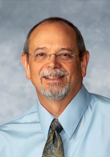 Headshot of Jon Fox, a middle-aged white man with rimless glasses, short hair and a light gray mustache and goatee. He's wearing a pale blue button-up shirt, a brown and blue paisley tie, and there's a gray marbled backdrop behind him. 