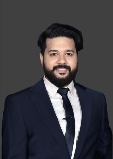 Headshot of Rahul Agrawal, a young man with dark black hair and a black mustache and beard. He's wearing a navy suit with a white shirt and navy tie. He's smiling at the camera and standing in front of a dark gray backdrop. 