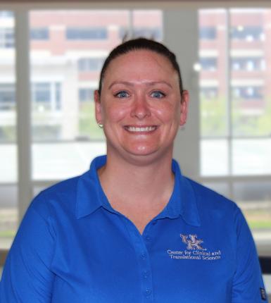 Headshot of Ellen Hartman, a white womane with blué eyes and dark hair pulled back tightly. She's smiling at the camera, wearing a blue polo shirt with the UK logo, and standing in front of a large window. 