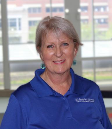 Headshot of Gail Sievert, a white woman with short gray hair. She's smiling at the camera, wearing a blue polo shirt and dangly blue earrings, and standing in front of a large window. 