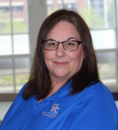 Headshot of Laura Ashe, a middle-aged white woman with dark straight hair just past her shoulders. She's smiling at the camera, wearing rectangular dark-rimmed glasses and a blue polo shirt, and standing in front of a large window. 