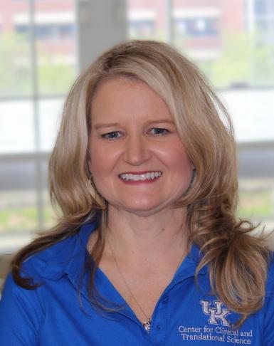 Headshot of a blond, middle-aged woman with hair past her shoulders. She's wearing a blue polo shirt with the UK logo on her left side. She's smiling at the camera and standing in front of a large window. 