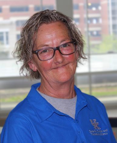 Haedshot of Terri Spear, a white woman with short, wavy gray hair and black plastic-rim glasses. She's smiling at the camera, wearing a blue polo shirt and standing in front a large window. 