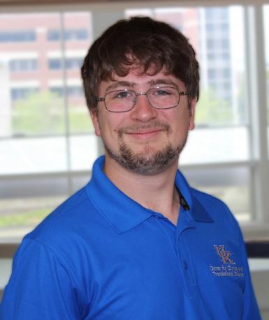 Headshot of Zachary Ross, a young white man with brown hair, a thin mustache and beard, and rectangular wire-framed glasses. He's smiling at the camera, wearing a blue polo shirt, and standing in front of a large window. 
