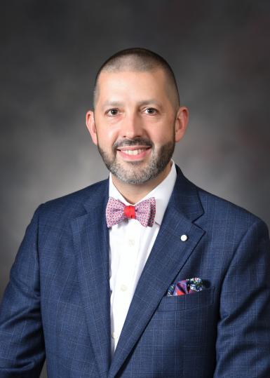 Headshot of Matt Bush, a white man with a buzz cut and short black and gray beard. He's smiling at the camera, wearing a blue suit jacket, red bow tie, and blue and red pocket square. A marbled gray backdrop is behind him. 