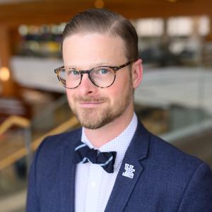 Headshot of Aaron Kruse-Diehr, a white man with short hair that's parted and slicked back; he's wearing tortoise-shell glasses, has a very short beard and mustache, and is wearing a dark blue suit jacket, light blue bow tie and a UK pin on his lapel. 