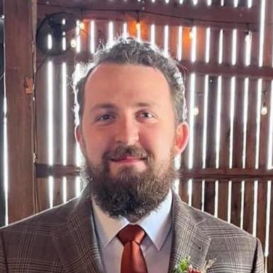 Headshot o Noah Perry, a white man with brown wavy hair pulled back and a brown mustache and beard. He's wearing a light brown plaid suit with a rust-colored tie. He's smiling at the camera, and behind him is a slatted wall through which sun is shining. 