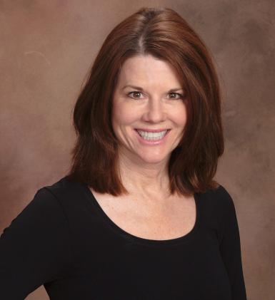 Professional headshot of Lynn Warneke, a white woman with straight, shoulder-length brown hair. She's wearing a black scoop-necked shirt and smiling at the camera. A brown photo backdrop is behind her. 