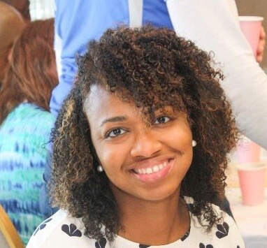 A young black woman with curly hair parted on the side and reaching just above her shoulders. She's smiling at the camera, wearing pearl stud earrings and a white blouse with black flowers.