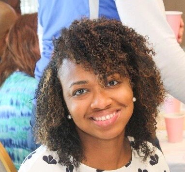 A young black woman with curly hair parted on the side and reaching just above her shoulders. She's smiling at the camera, wearing pearl stud earrings and a white blouse with black flowers. 