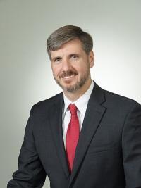 Professional studio photo of Kevin Hatton, a thin, middle-aged white man wearing a dark gray suit, white collared shirt, and red tie. He has light brown/gray hair parted on the side, and as short beard and mustache. He's smiling at the camera. 