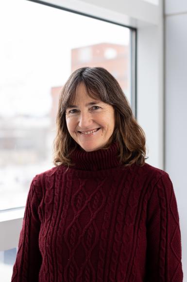 Photo of Jennifer Stevenson, a middle-aged white woman with shoulder length, wavy brown hair. She's smiling at the camera and wearing a dark red cable-knit turtleneck sweater. She's standing in front of a window. 