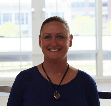 A young white woman in a dark shirt smiles at the camera in front of a wall of windows. Her hair is slicked back into a bun and she's wearing a necklace with a large stone. 