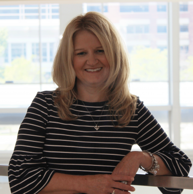 A middle-aged white woman with dark blond hair past her shoulders leans against a metal railing and smiles at the camera. She's wearing a navy shirt with white stripes. Behind her is a wall of windows. 
