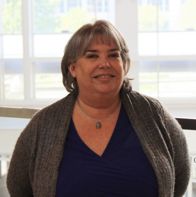 A middle-aged white woman with shoulder length brown-gray hair smiles at the camera. She's wearing a gray cardigan, navy shirt, and a necklace with a small charm. Behind her is a wall of windows.  