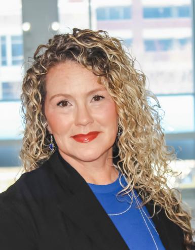 Photo of a white woman with long, curly blond hair. She's giving a calm smile to the camera and wearing red lipstick, a black blazer, and a royal blue shirt. Behind her are windows. 