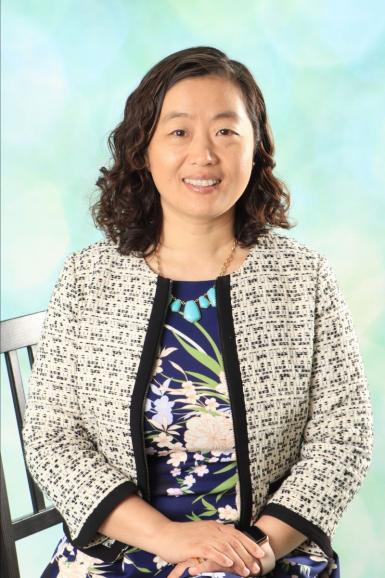 Photo of an Asian woman with shoulder-length wavy hair. She's sitting in a chair with hands folded in her lap. She's wearing a cream-colored blazer and navy flowerful dress, and smiling at the camera. 