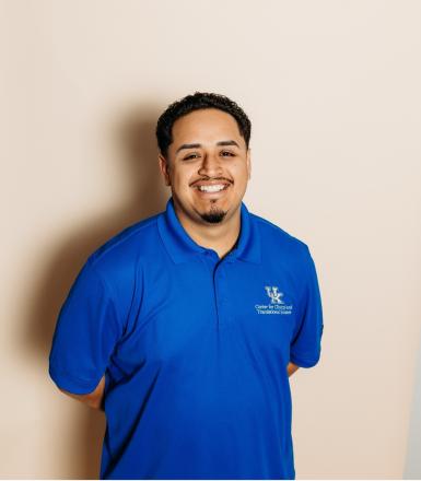 Photo of a young man smiling at the camera. He has black hair, a thin black mustache and goatee, and is wearing a royal blue polo shirt with the UK logo on the right side. 