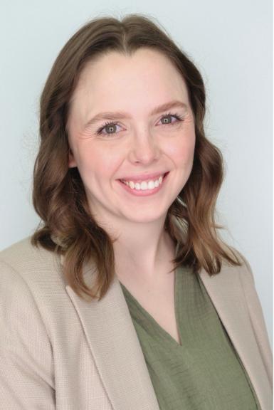Headshot of a young white woman with wavy light brown hair just past her shoulders. She's smiling at the camera, wearing a tan blazer and a green blouse. 