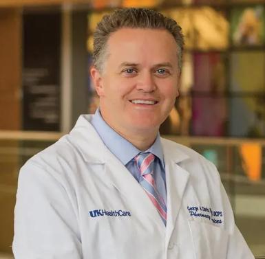 Indoor portrait photo of a middle-aged white man wearing a white lab coat, light blue collared shirt, and light blue and pink striped tie. He has short gray hair, blue eyes, and is smiling at the camera. 
