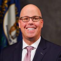 Man with a bald head and rectangular glasses smiles at camera wearing a dark blue suit, white shirt and light pink tie