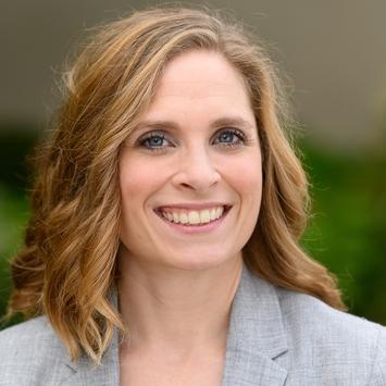 Headshot of a woman with dark blonde shoulder-length hair, wearing a light gray suit jacket and smiling. 